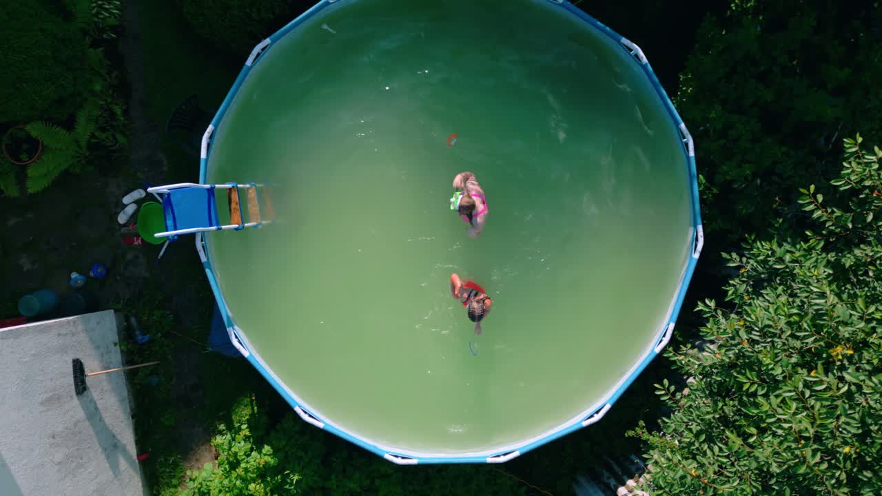 Overhead view of two people swimming in green circular backyard pool surrounded by lush garden trees