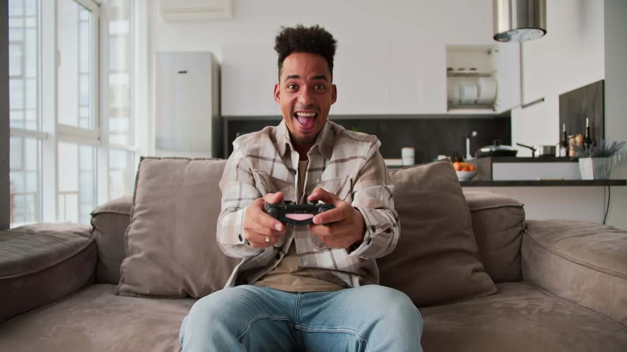 Portrait of a happy young Black-skinned brunette man with stubble who sits on a modern sofa and plays a video game and rejoices at his victory using a black joystick in a modern studio apartment