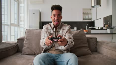 Portrait-of-a-happy-young-Black-skinned-brunette-man-with-stubble-who-sits-on-a-modern-sofa-and-plays-a-video-game-and-rejoices-at-his-victory-using-a-black-joystick-in-a-modern-studio-apartment