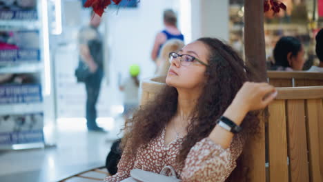 dama con cabello rizado, vestido vintage, y gafas sentada en un banco de madera, ajustando el cabello con expresión reflexiva, el fondo muestra a personas de pie y sentadas con luz suave creando un ambiente cálido
