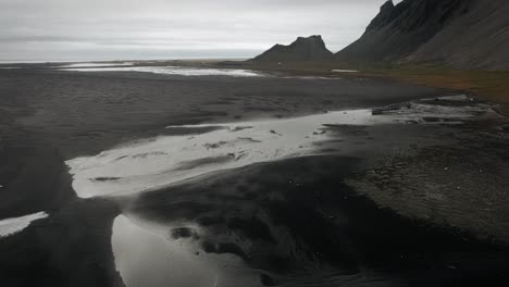 Schwarzer-Sandstrand-Aus-Der-Luft,-Stokksnes,-Reflexionen-Vulkanischer-Dunkler-Berge-In-Der-Ferne,-Düstere,-Stimmungsvolle,-Wolkige-Landschaft-Islands