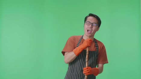 ama de llaves masculina asiática con un delantal cantando a la escoba de barrer mientras está de pie en el estudio de pantalla verde