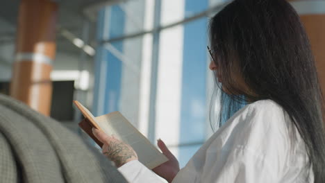 side view of lady with floral tattoo flipping to new page while reading book, standing near modern glass railing with checker-pattern cloth resting on rail in bright urban interior