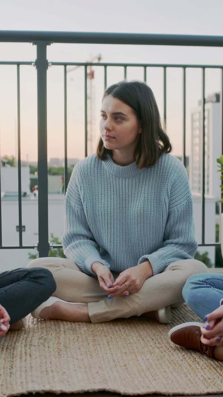 Vertikal video: Lytter til ven, kvinde i blå sweater vender sig, gestikulerer på tagtæppe med telefon.