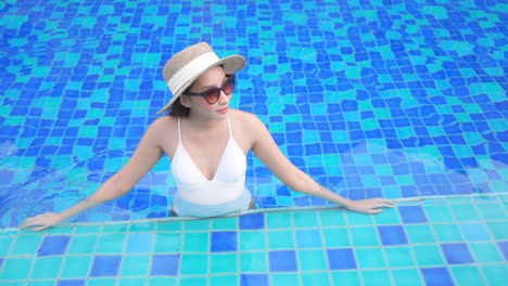 Elegante-Dama-Asiática-En-Piscina-Azul-Con-Sombrero-De-Verano-Disfrutando-De-Vacaciones-Tropicales,-Cámara-Lenta