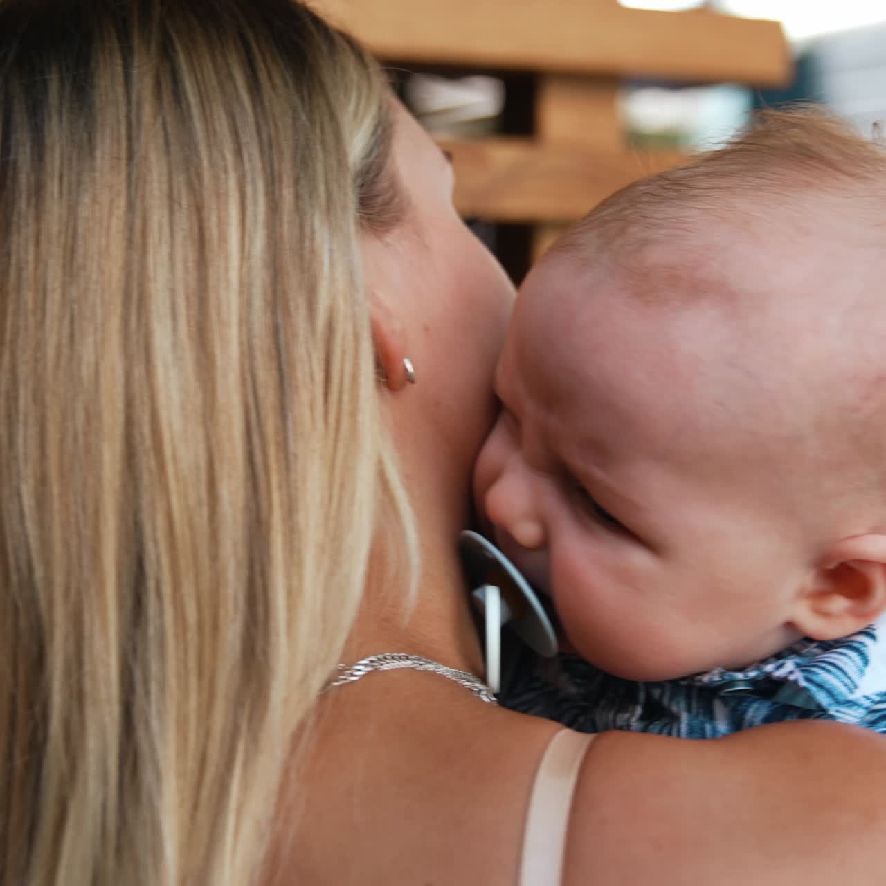 Blonde woman is holding her baby boy. Mother is calming her infant with pacifier in mouth. Close up