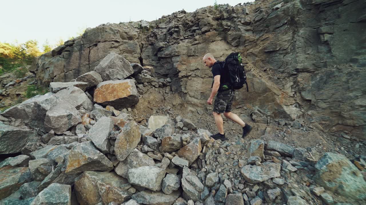 extreme tourist in a black t-shirt and shorts with a backpack on his shoulders is walking along the rocks and climbing a large stone in a warm weather in the summer