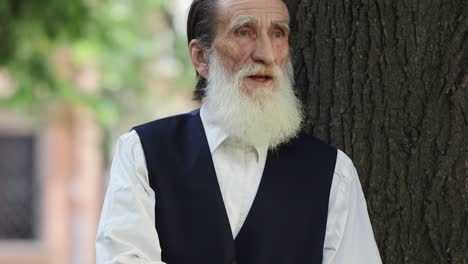 portrait of an elderly man with a beard leaning against a tree