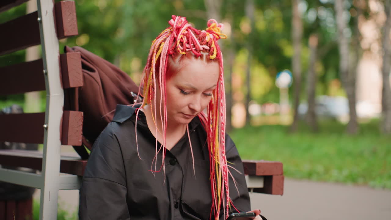 Mujer blanca con rastas rosas, auriculares, desplazándose por una lista de reproducción y cerrando los ojos brevemente mientras está sentada en un banco, luz solar moteada entre los árboles, componente visual del audio como herramienta de afrontamiento y consuelo personal