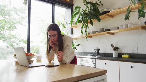 mujer transgénero caucásica no binaria que usa computadora portátil y teléfono inteligente en la cocina