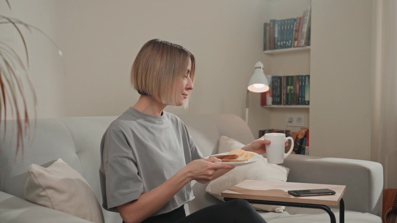 Scena di colazione accogliente sul divano con una donna caucasica che porta un vassoio al divano, posizionando tazza e toast, sistemandosi per mangiare mentre guarda il telefono per la musica, calda libreria e lampada sullo sfondo, mattina domestica.