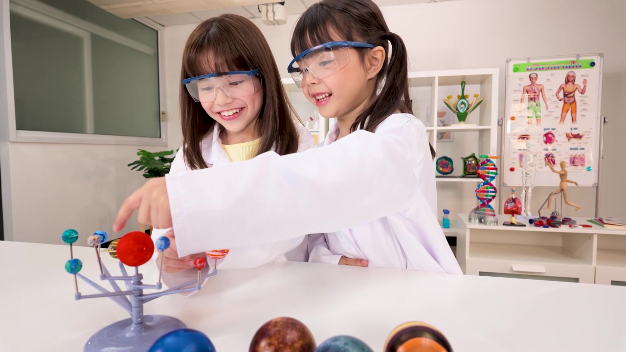 Two young students in lab coats and goggles examine a planetary model during science class.