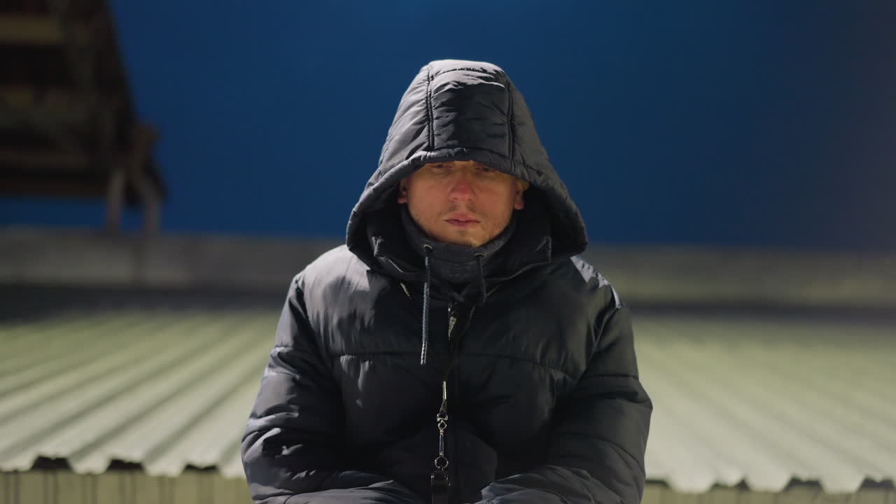 검은 옷과 후드 재을 입은 남자가 다리 사이에 손을 고 혼자 앉아있고, 냉정하게 보이며, 그는 희미하게 밝은 배경으로 경기장에서 있습니다.