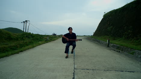 young asian male sitting in the middle of a road playing a guitar