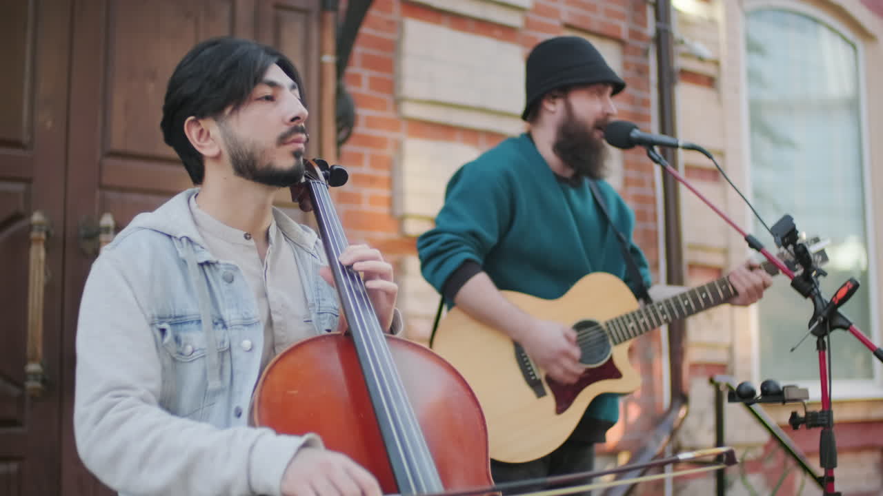 Sequence of shots of man playing cello and male vocalist with acoustic guitar singing into microphone while performing outside for passerbys