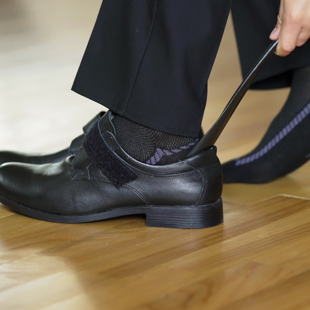 Teens legs are putting on new fashion shoes with the help of a shoe spoon on parquet. A boy in black socks puts on school shoes on the brown floor background indoors at home.