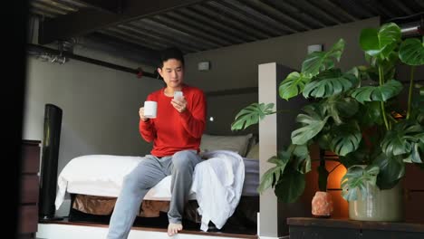 Animation-of-network-of-digital-icons-over-asian-man-with-coffee-using-smartphone-on-bed-at-home