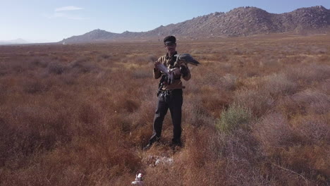 young man training falcon stands in dry brown brush desert with hawk