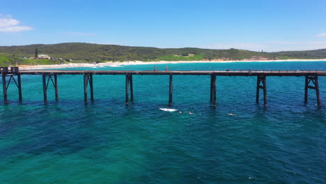 Young-male-adult-jumping-off-pier-into-sea,-aerial-pull-away-reveal