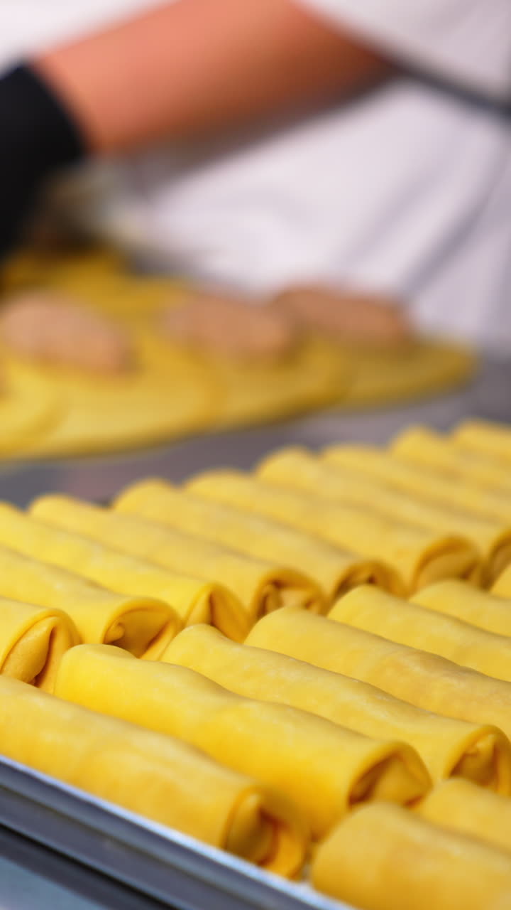 Full tray with semi-cooked stuffed crepes. Close up. Chef in gloves rolls yellow pancakes with stuffing at backdrop in blur. Vertical video