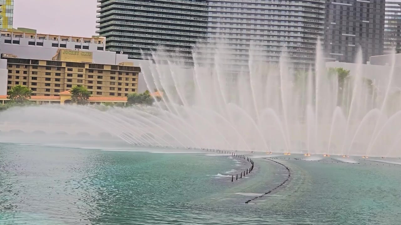 Vista amplia de chorros de fuentes sincronizadas en Las Vegas, rociando sobre un lago de resort turquesa con hoteles altos, arcos de piedra, vegetación, ondas y un brillante paisaje urbano.