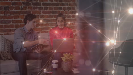 animation of glowing network over male and female colleague using laptop on couch in office