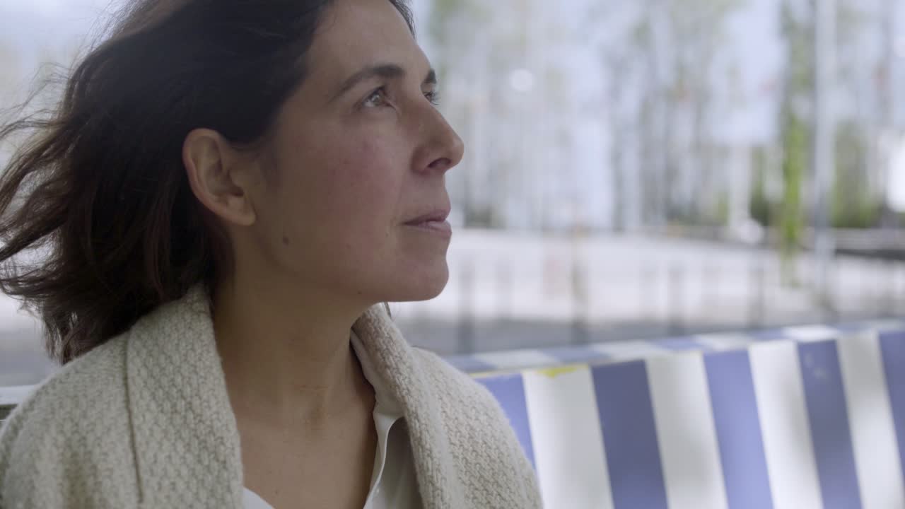 vista lateral de una atractiva morena sentada en un banco a rayas al aire libre.