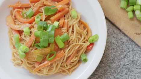 fideos de espagueti con pimientos cocidos en rodajas de colores, zanahorias y puerro fresco con salsa de maní en un plato blanco al vapor con más puerro en rodajas en una tabla de madera