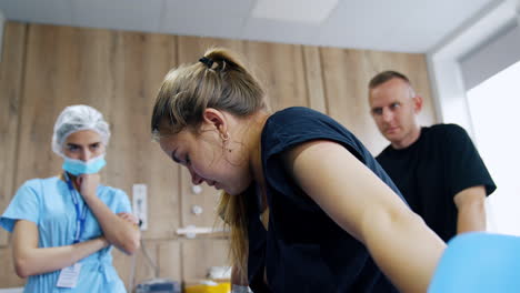 giving birth to a baby. blonde caucasian woman stands near the bed. husband and uniformed medic at backdrop.