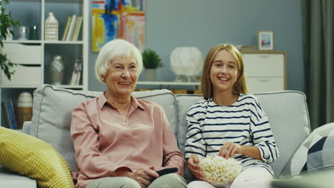 portrait shot of the pretty small teenager girl and senior grandmother sitting together on the sofa at home, watching tv and laughing