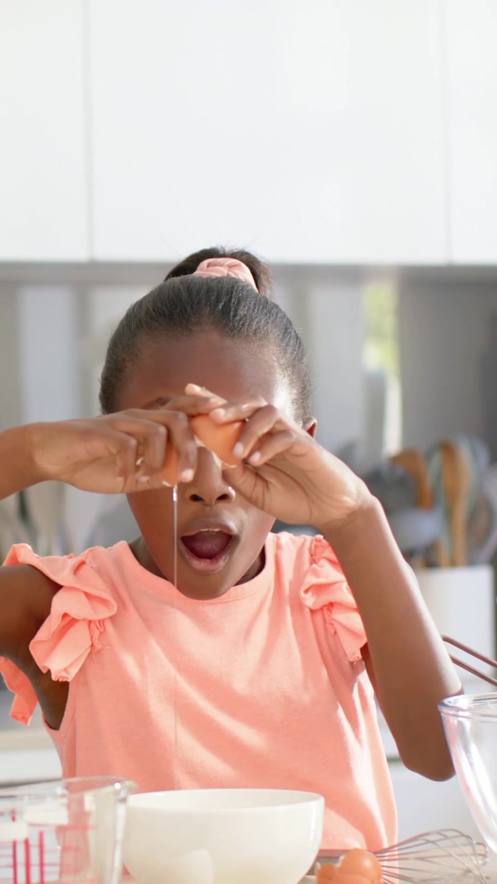 Vertical video: Lifting egg, girl in pink top cracking into bowl in kitchen making batter