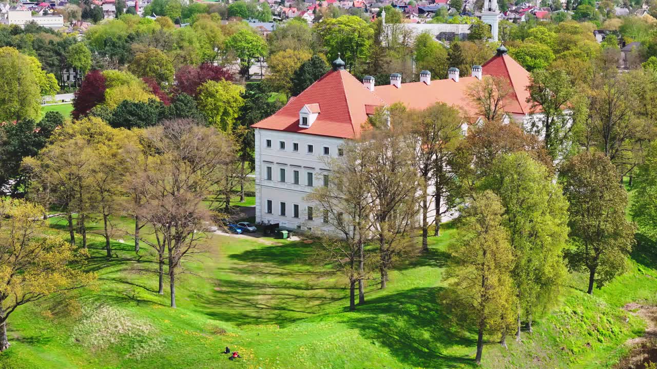 A striking Baroque-style manor with a red tile roof rises above a lush, sloped hill, surrounded by leafing spring trees and distant village rooftops.