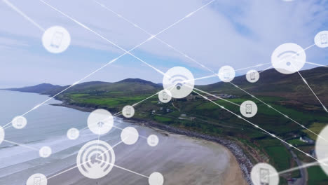 animation of network of digital icons against aerial view of landscape with mountains, beach and sea