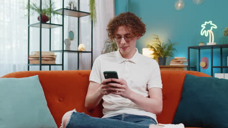 Caucasian-young-man-sitting-on-couch-using-mobile-smartphone-share-messages-smiles-at-home-room