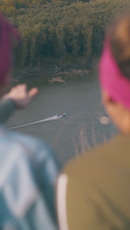 girl tourists look at sailing motorboat from steep river bank in warm autumn evening close backside view