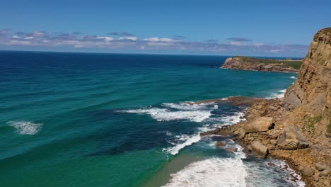 Drone-flight-in-reverse-over-the-cliff-coast-of-orange-rock-and-grass-on-a-sunny-summer-day-with-calm-sea-and-a-horizon-of-clear-blue-sky-over-the-turquoise-and-blue-sea-in-Cantabria-Spain