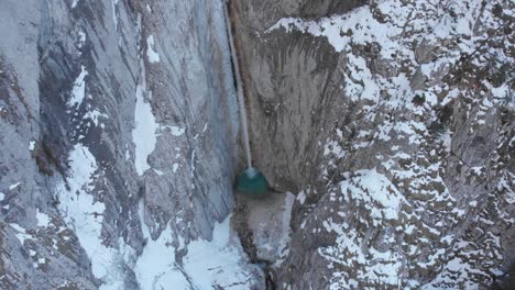 Dron-flying-up-looking-down-on-the-waterfall-with-the-rock-wall-covered-in-snow-in-the-winter