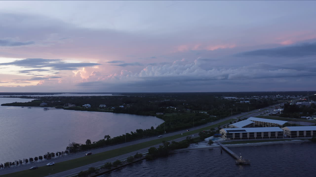 A quiet coastal city stretches beside calm bay waters as dusk settles, where soft light glows across storm clouds gathering over the horizon in shades of violet and rose