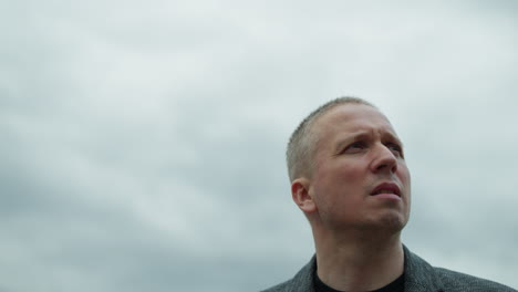 a close-up view of an aged white man wearing a grey suit and a black polo shirt, looking around with a serious expression