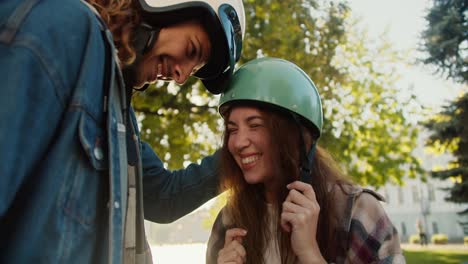 Nahaufnahme-Eines-Glücklichen-Brünetten-Mädchens-In-Einem-Grünen-Mopedhelm-Und-Einem-Karierten-Hemd,-Das-Ihren-Freund-Mit-Lockigem-Haar-In-Einer-Jeansjacke-In-Einem-Städtischen-Sommerpark-Umarmt-Und-Ansieht