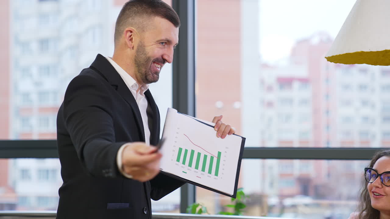 Male mid-aged boss stands in front of his team and shows a document. Businessman explaining chart to his office staff at meeting.