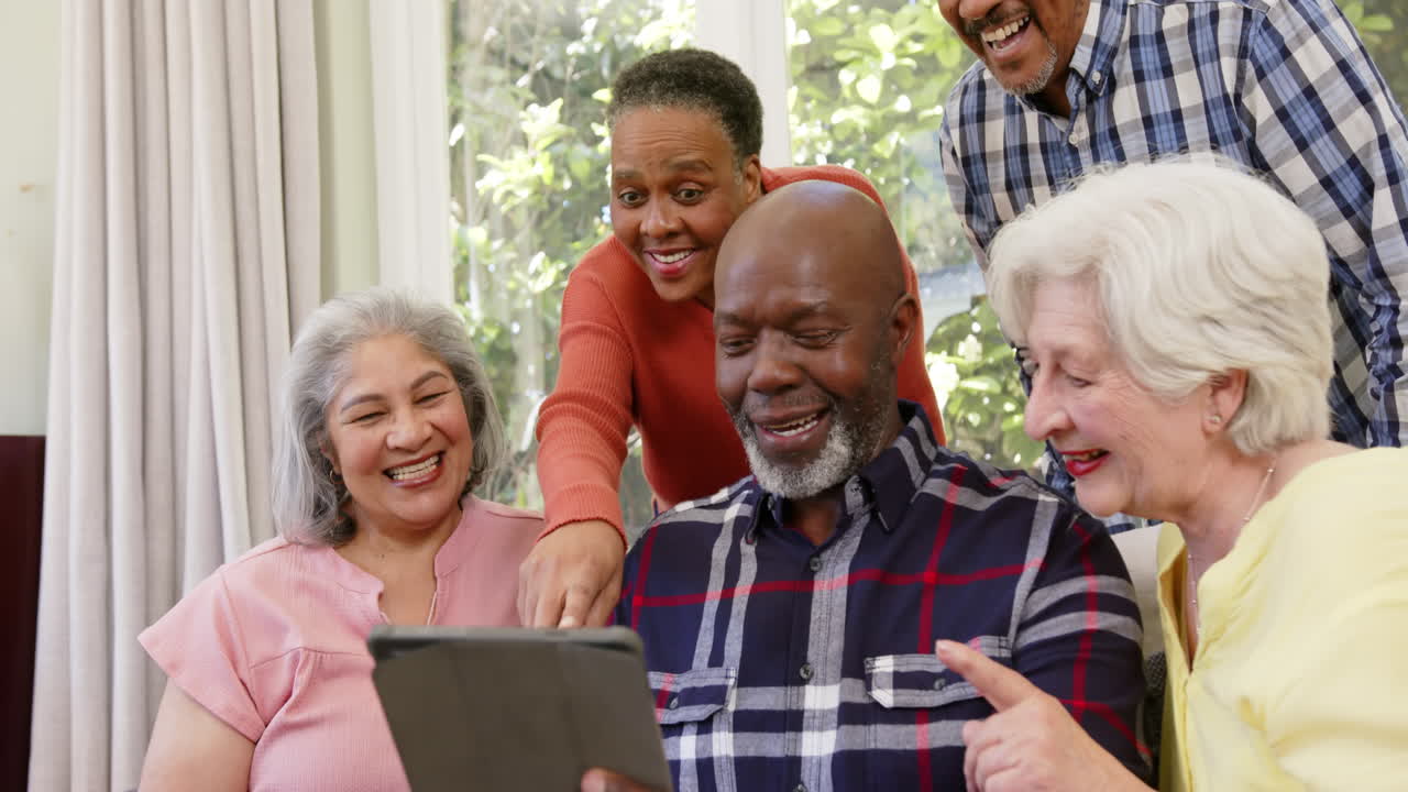 Happy diverse senior female and male friends looking at tablet together in sunny room, slow motion