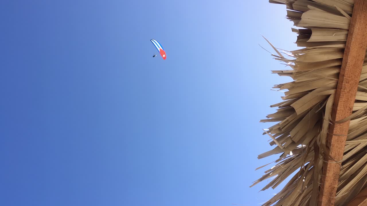 paraguas de playa de paja, cielo azul y persona en parapente con ala de bandera cubana