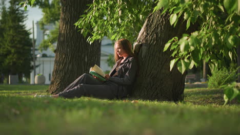 woman sitting outdoors with legs stretched, leaning against a tree on a grassy field, flipping through pages of a book, tree leaves swaying gently in the breeze, and greenery in the background