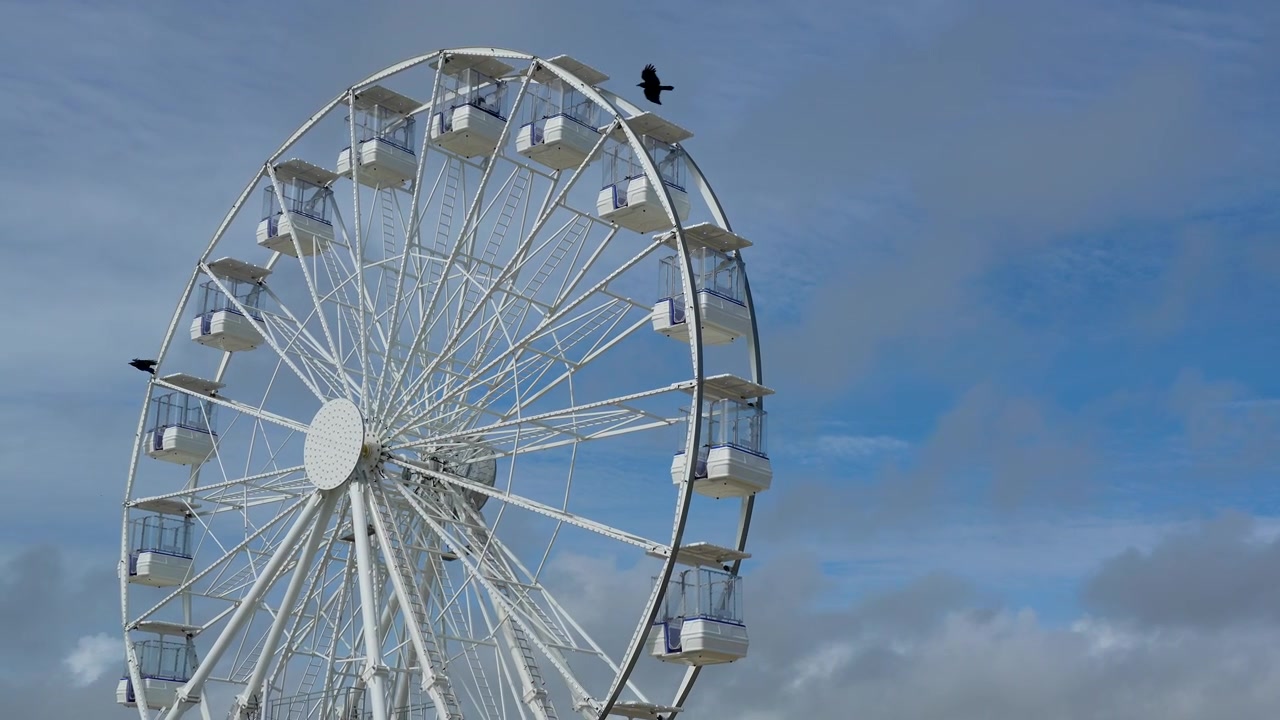 Una grande ruota panoramica bianca che gira lentamente in una località balneare. Uccelli volano oltre l'attrazione sotto un cielo azzurro brillante con nuvole soffici