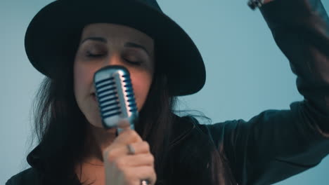 a singer in a black hat and a black leather jacket holds a vintage microphone with one hand while gesturing with the other, set against a blue background