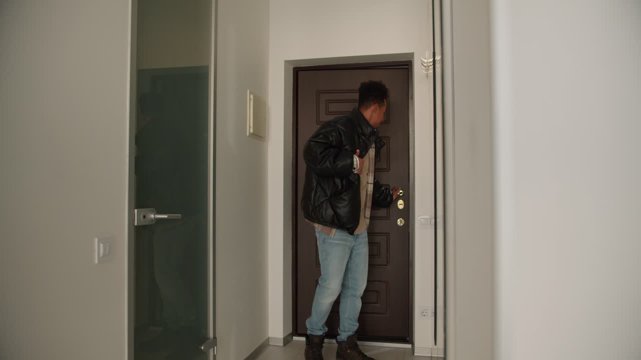 un joven feliz con piel negra abre la puerta principal de la casa y se va a casa después de lo cual se quita su ropa exterior y entra en su casa. joven feliz viene a casa con una chaqueta negra y se la quita mientras camina dentro. con una camisa beige a cuadros
