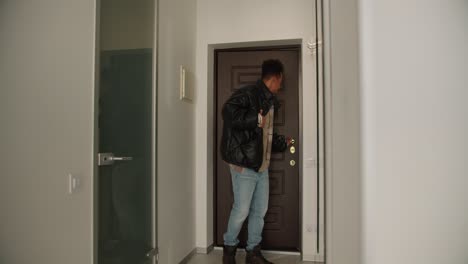 A-young-happy-man-with-Black-skin-opens-the-front-door-of-the-house-and-goes-home-after-which-he-takes-off-his-outerwear-and-goes-into-his-house.-Happy-young-man-comes-home-wearing-a-black-jacket-and-takes-it-off-while-walking-inside.-Wearing-a-checkered-beige-shirt