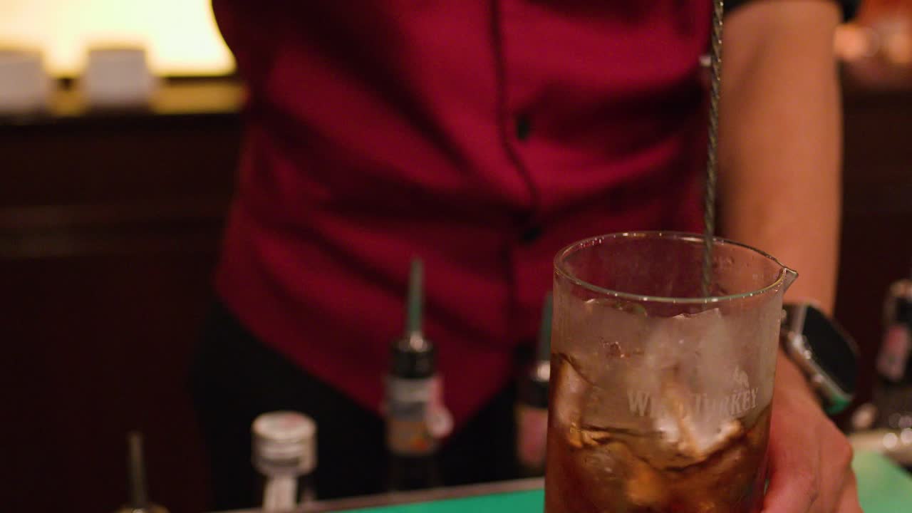 Bartender stirs whiskey cocktail with bar spoon in dimly lit Bangkok nightlife bar setting
