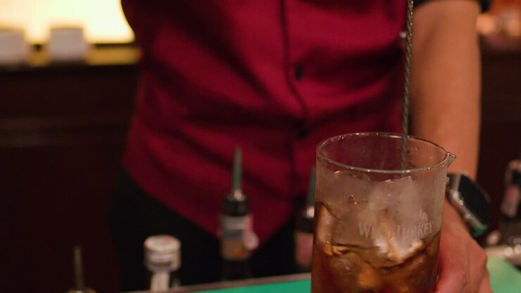 Un barista mescola un cocktail a base di whisky con un cucchiaio da bar in un bar notturno scarsamente illuminato a Bangkok